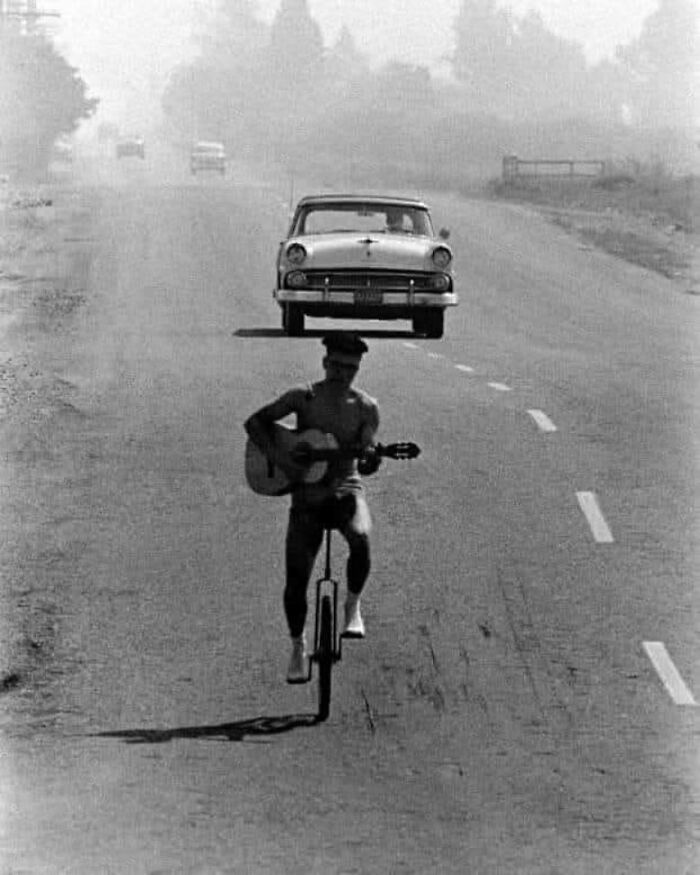 Man plays guitar on unicycle with vintage car behind, capturing a forgotten corner of history.