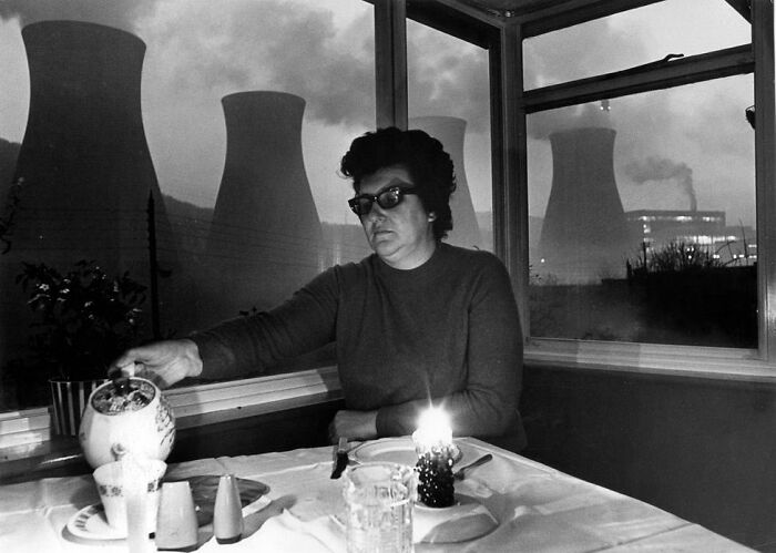 A woman pours tea at a table, silhouetted by cooling towers, capturing a forgotten corner of history.