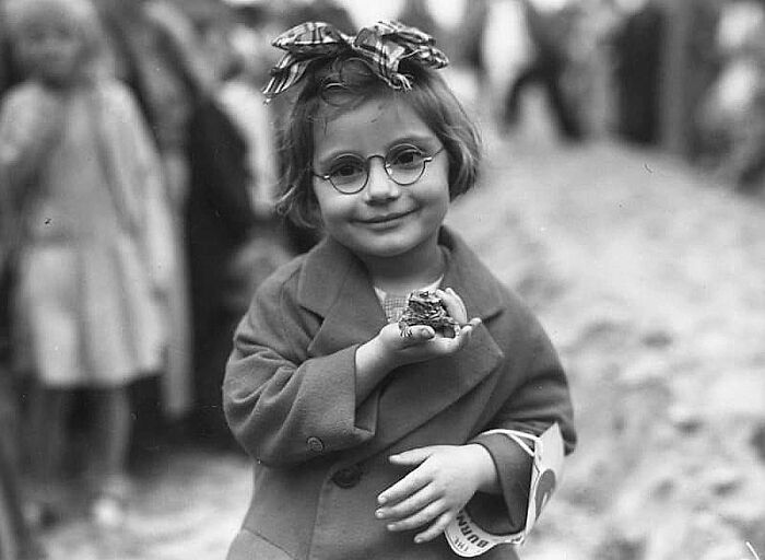 Young girl with glasses, wearing a coat and ribbon, holding a small object, in a historical setting. Historical photo featured.