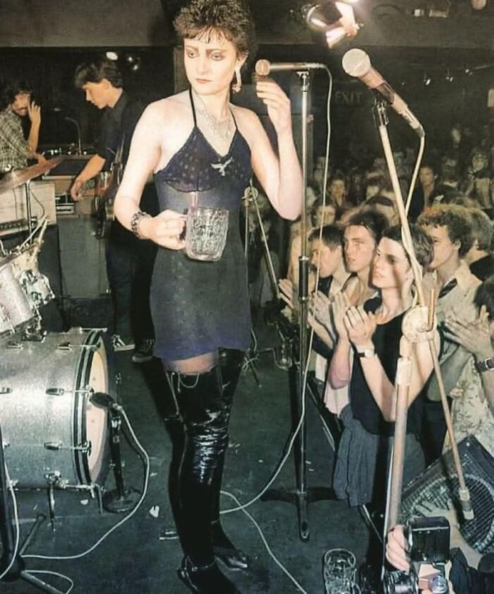 Musician performing on stage in black boots, holding a drink, with audience clapping; a glimpse into forgotten corners of history.