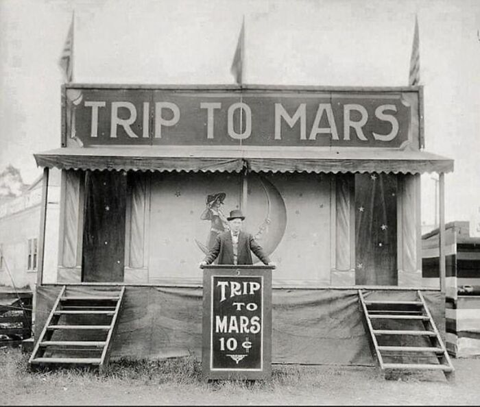 Man in front of a vintage "Trip to Mars" carnival booth, offering tickets for 10 cents.