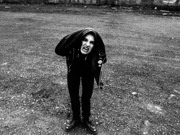 Person in leather jacket, hood up, making a fierce expression in a gravel lot. Forgotten corners of history.