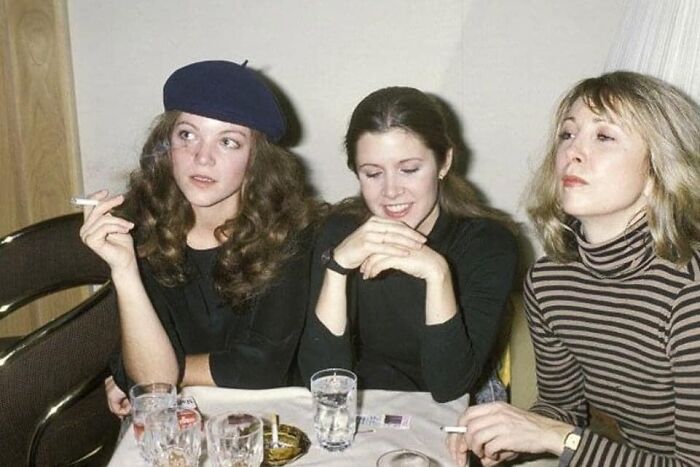 Three women from forgotten corners of history, sitting at a table with drinks and cigarettes, in casual attire.