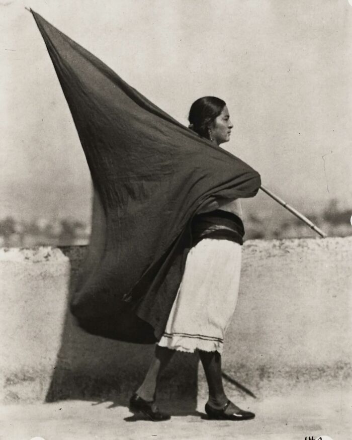 A woman standing on a rooftop with a large flag, a scene from forgotten corners of history captured in black and white.