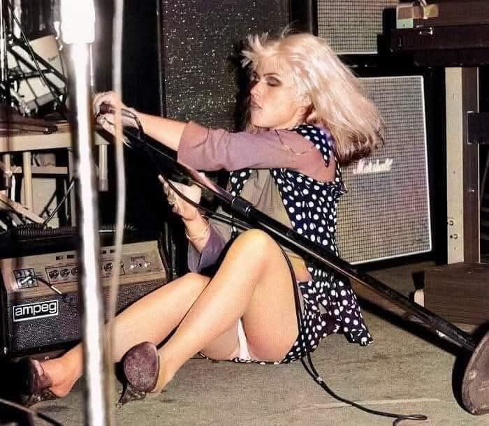 Blonde woman sitting on a stage during a performance with a microphone stand, capturing a forgotten corner of history.