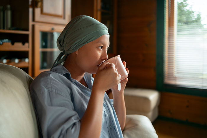 Employee with cancer diagnosis, wearing headscarf, sips from a mug on a cozy couch by a window. Employee with cancer diagnosis, wearing headscarf, sips from a mug on a cozy couch by a window.