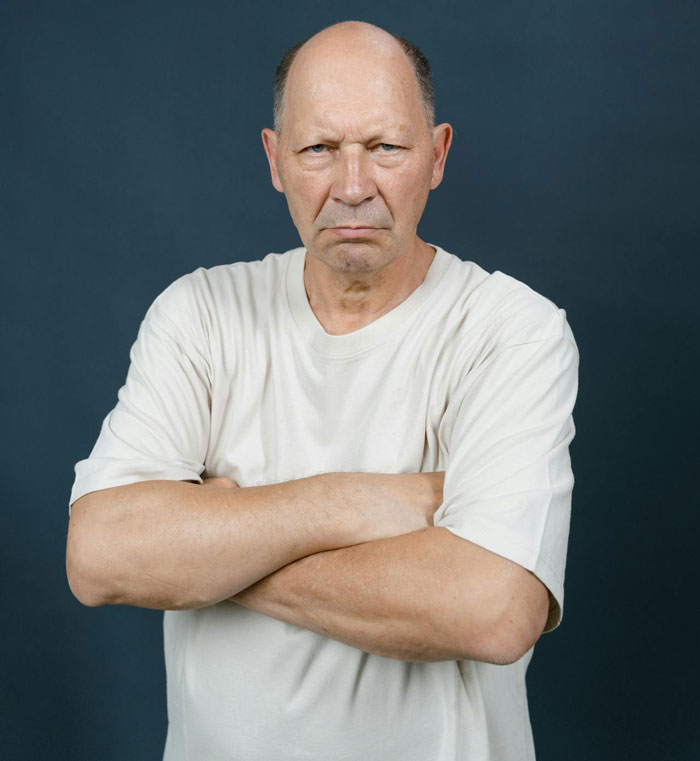 Elderly man in a white t-shirt with arms crossed, looking displeased, representing homophobic family reaction.