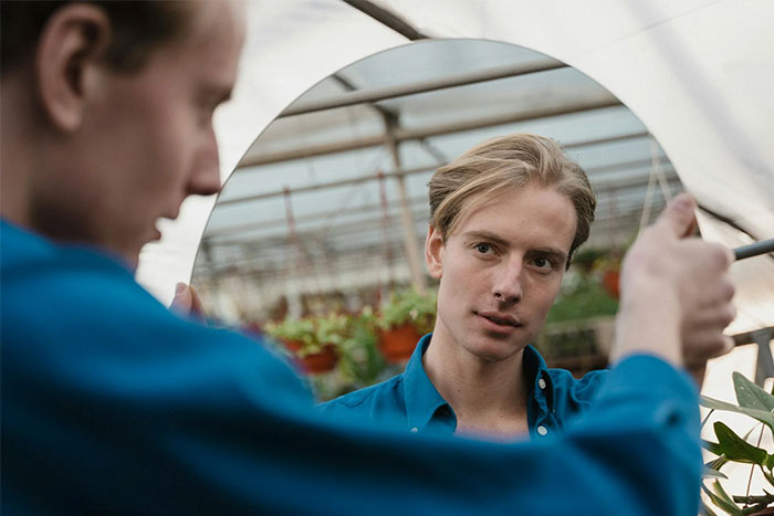 Man in a blue shirt looking at his reflection in a round mirror, inside a greenhouse.