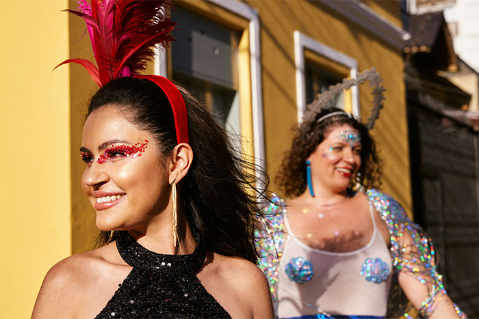 Two women in colorful costumes and makeup smiling, with one wearing a red feathered headpiece.