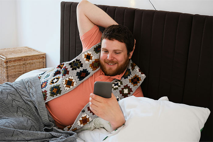 Man lying on bed, smiling at phone, cozy setting, colorful blanket.