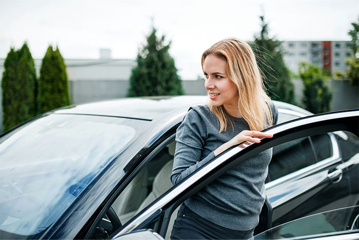 A woman in a gray sweater smiling as she stands by her car, with trees and buildings in the background.