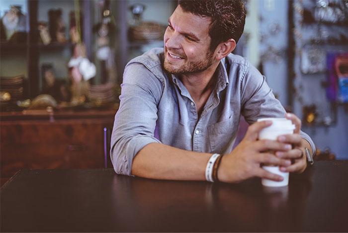 Man in casual shirt smiling and holding a coffee cup at a table; related to women's dating experiences with men.