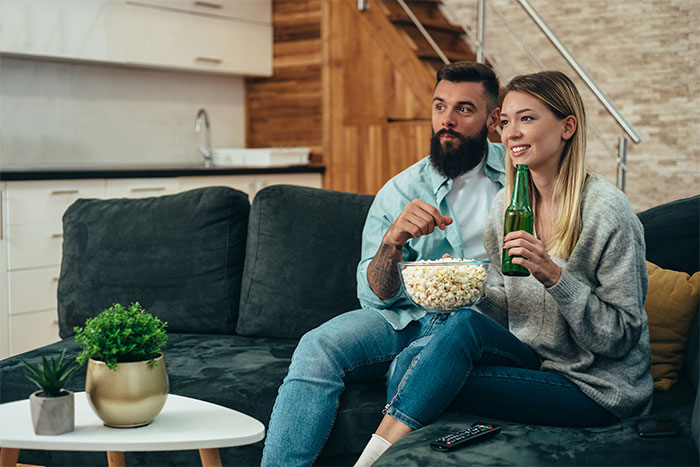 Couple on couch watching TV, woman holding a beer, man with popcorn, illustrating dating experiences.