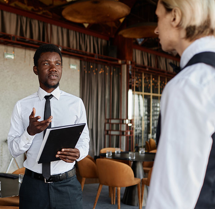 Two employees in uniform talking in a restaurant setting, holding a clipboard.