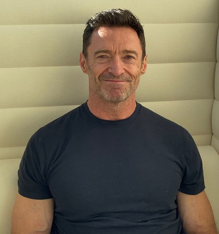 Hugh Jackman in a casual setting, seated and smiling, wearing a dark t-shirt.