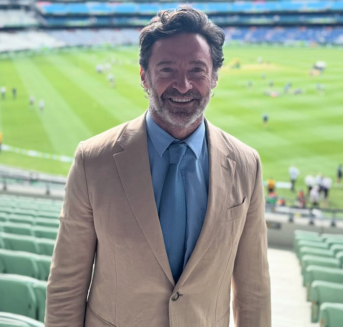 Person in a beige suit and blue tie at a sports stadium, showcasing a new facial look, aligning with the 70s Pawn Star style.