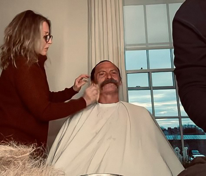 Man receives a 70s-inspired facial makeover while sitting in chair, woman adjusts mustache in well-lit room.