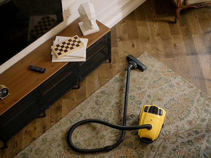 Yellow vacuum cleaner on a floral rug next to a TV stand with books and a chessboard; husband gift idea.