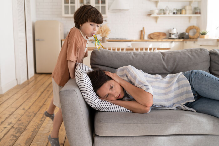 Mother in despair on couch, toddler playing nearby in a living room setting, reflecting parenting challenges.