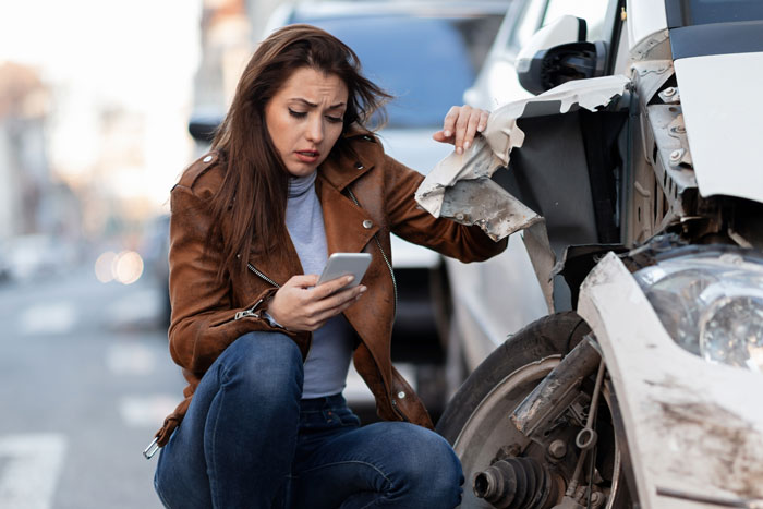 Woman in a brown jacket, looking concerned at her phone beside a damaged car, representing financial stress.
