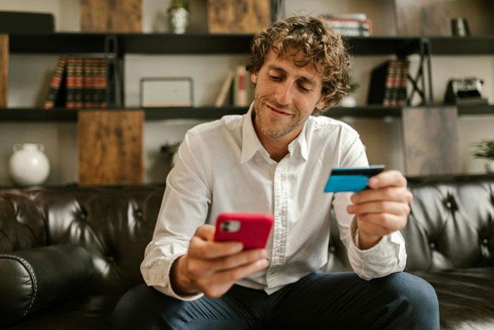 Man in white shirt on sofa holding phone and credit card, illustrating a secret $7.5K loan situation.