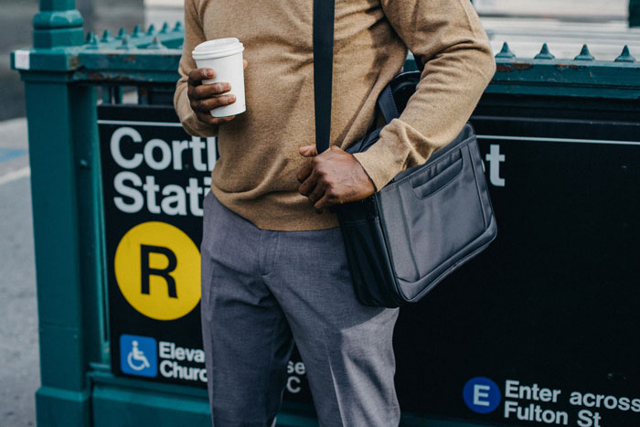 Man holding coffee cup at Cortlandt Street Station entrance, wearing a brown sweater and gray pants, with a black bag.