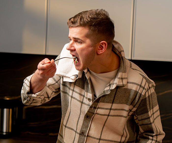 Man eating in kitchen, wearing a plaid shirt; related to lunch being called "absolutely disgusting. Man eating in kitchen, wearing a plaid shirt; related to lunch being called "absolutely disgusting.