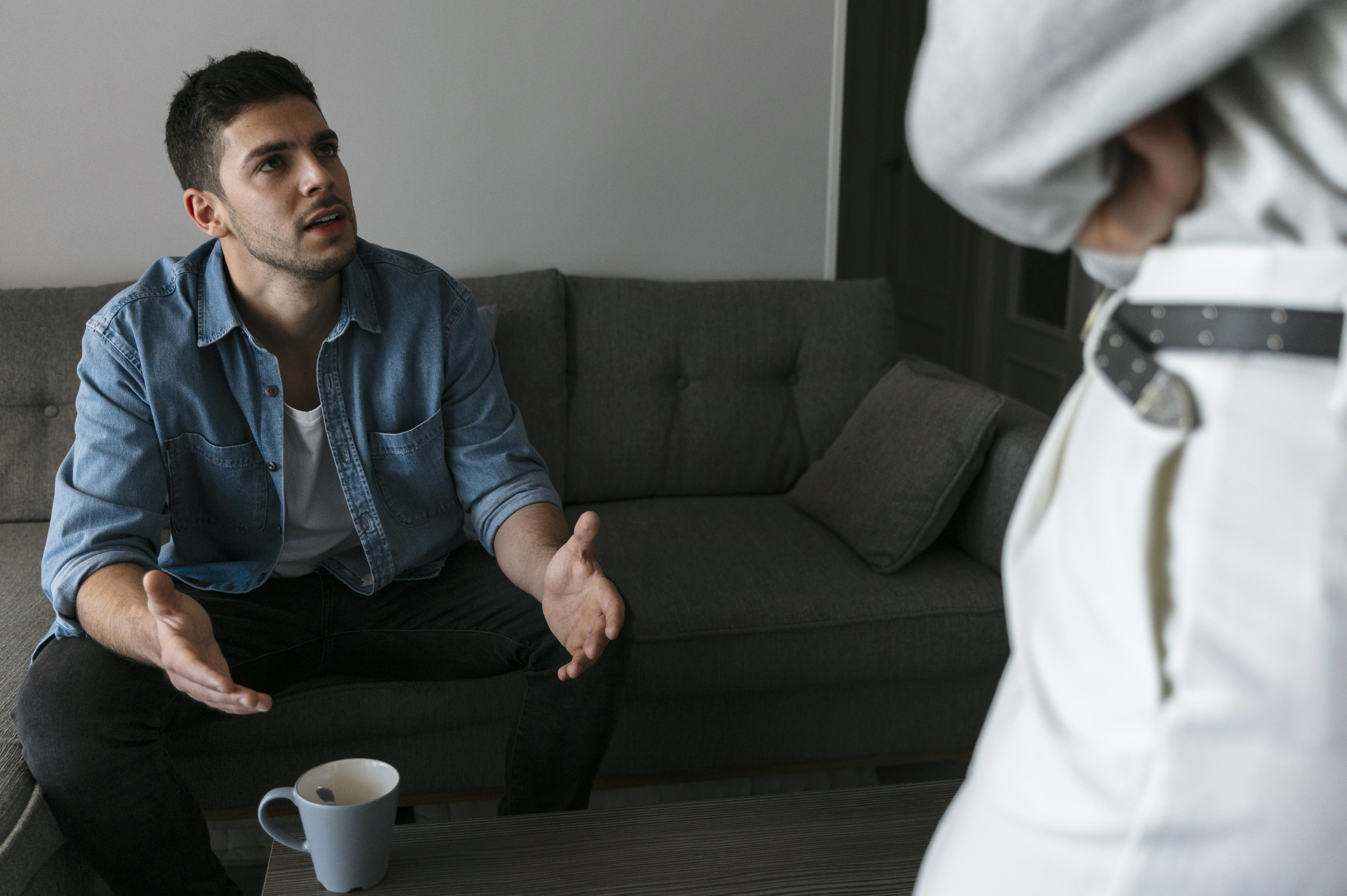 Man gesturing while seated on a couch, with a coffee cup on the table in a domestic setting.