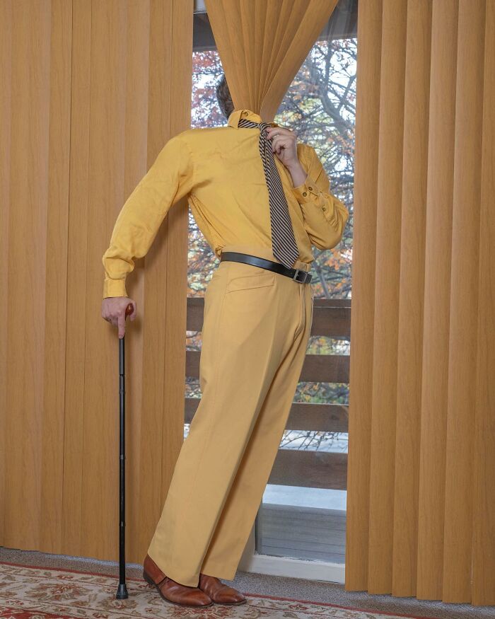 Solitary figure in a yellow suit blending into curtains, holding a cane, in a surreal photograph by Ben Zank.