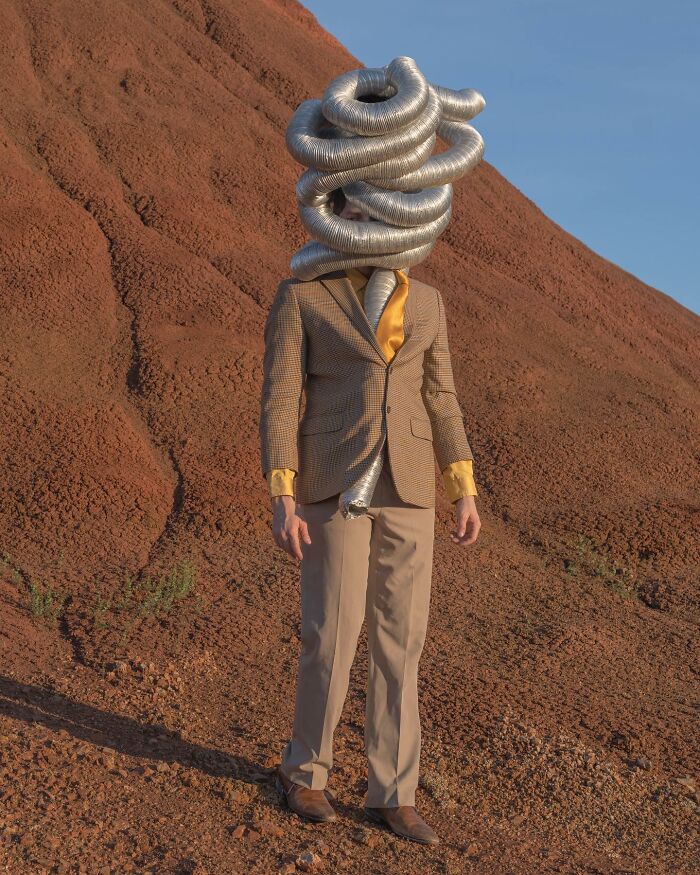 A figure in a suit with a coiled metal duct headpiece stands in a surreal desert landscape by Ben Zank.