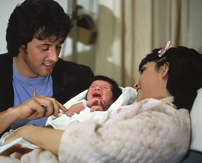 A Sylvester Stallone and Talia Shire, cuddles a newborn baby, capturing a tender family moment away from fame.