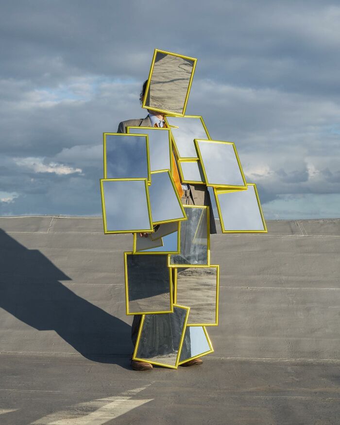 Solitary figure covered in mirrors on a rooftop under a cloudy sky, creating a surreal reflection of the surroundings.
