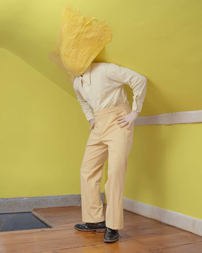 A surreal photograph by Ben Zank featuring a solitary figure with a yellow paper covering their head in a yellow room.