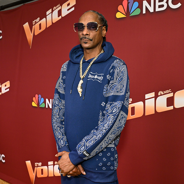 Snoop Dogg in blue bandana-pattern hoodie and sunglasses, standing on "The Voice" red carpet, representing rap royalty and business.