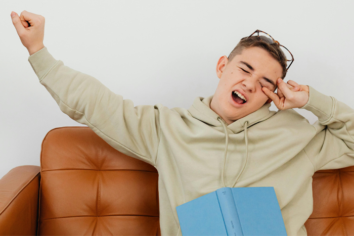 Young person yawning, sitting with a book on a couch, representing boredom and looking for ways to entertain themselves.