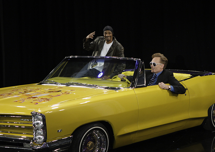 Conan O'Brien posing next to Snoop Dogg in sunglasses driving a yellow car, symbolizing rap royalty and high-stakes lifestyle.