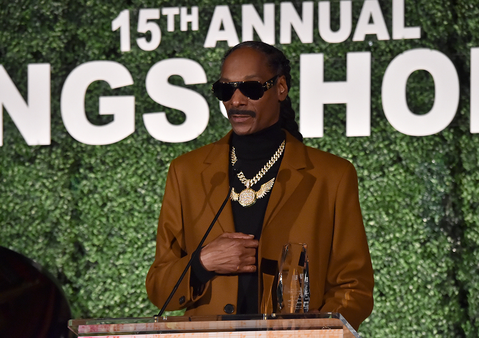 Snoop Dogg in sunglasses and gold chain, speaking at an event, representing rap royalty and business success.