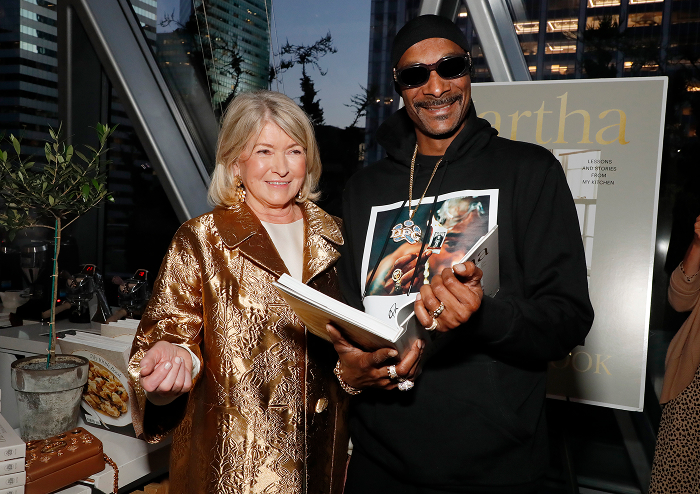 Snoop Dogg holding a book, smiling alongside a woman in a gold coat, highlighting his shift from rap royalty to business deals.