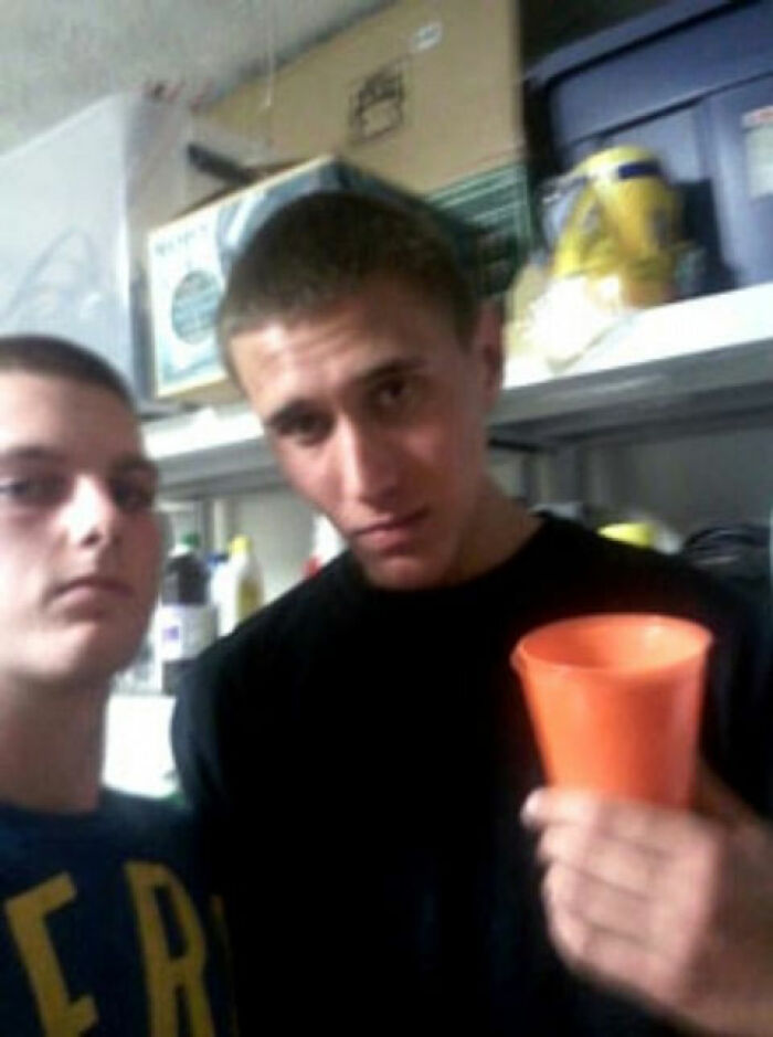 Two young men in a dimly lit room, one holding an orange cup, surrounded by storage containers. Creepy photo context.