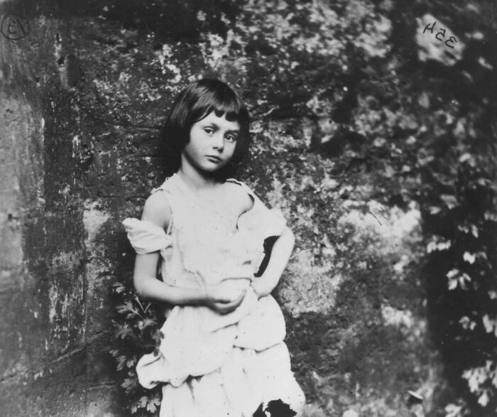 Young girl in an old photograph, wearing a tattered dress, standing against a stone wall, evoking a sense of creepy history.