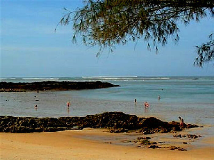Beach scene with dark history, showing scattered people in shallow water, rocky shore, and distant waves under a clear sky.