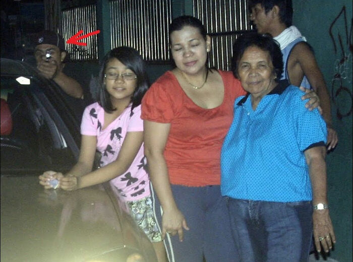 Family posing in front of a car while a figure in the background with a camera adds an eerie element to the photo.