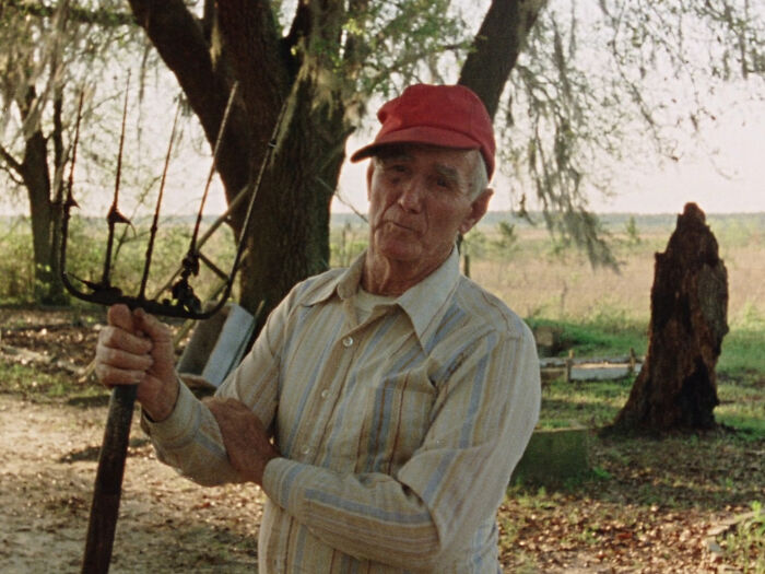 Elderly man in a striped shirt and red cap holding a pitchfork in a rural setting.