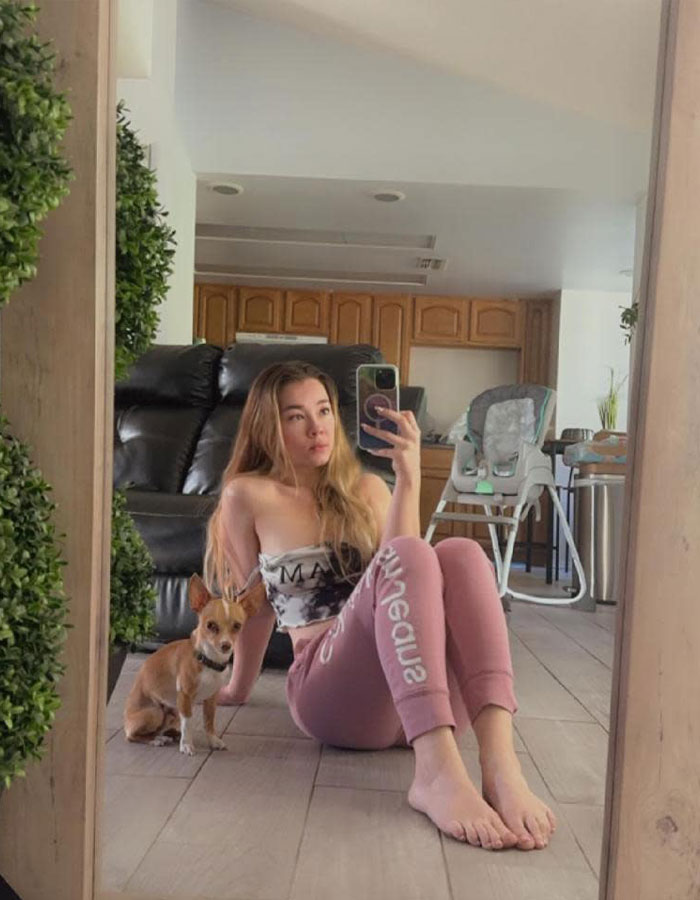 Influencer sits on floor with dog, taking a mirror selfie in a living room setting.
