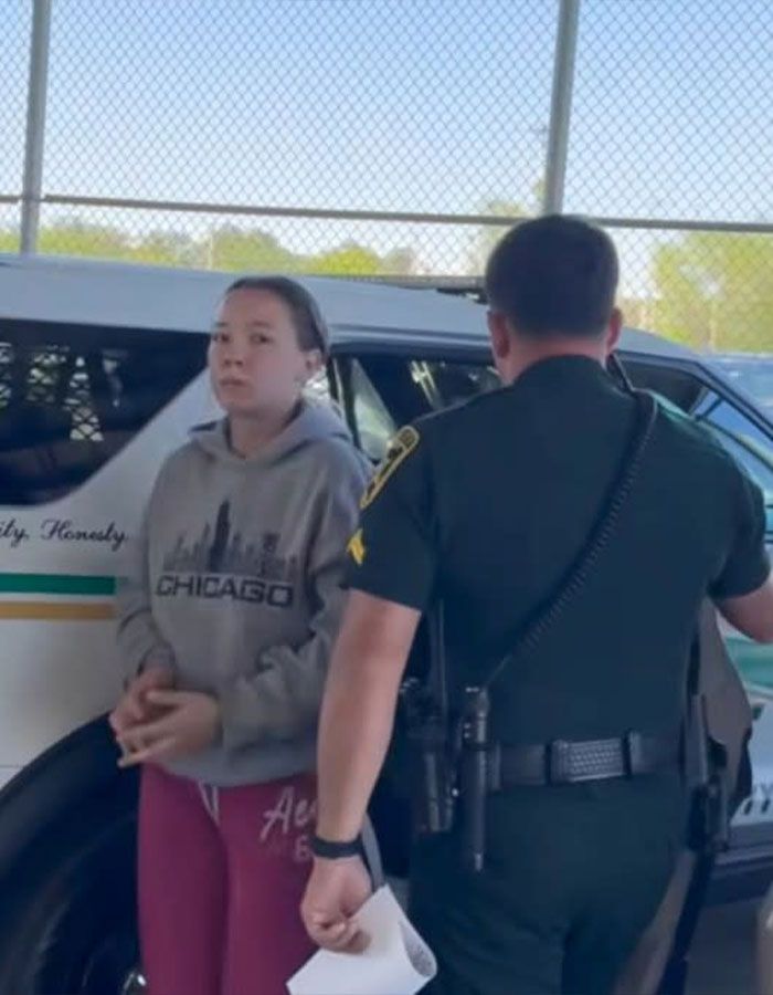Influencer arrested near police car, wearing a gray Chicago hoodie, escorted by an officer in a green uniform.