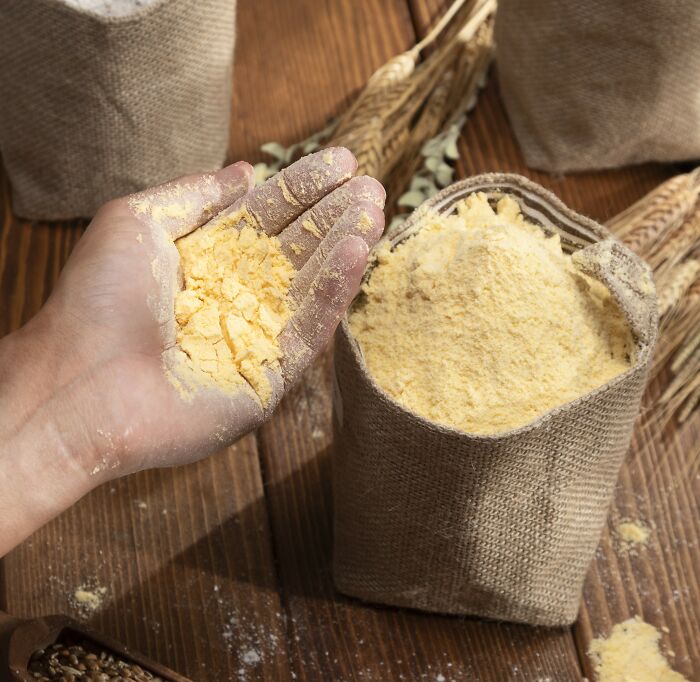 Hand holding cornmeal next to a burlap sack on a wooden table, illustrating homemade bread ingredients.