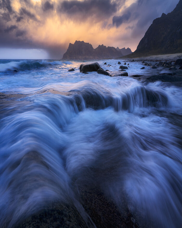 Stunning landscape of waves crashing on rocky shore at sunset, with dramatic clouds and distant mountains.