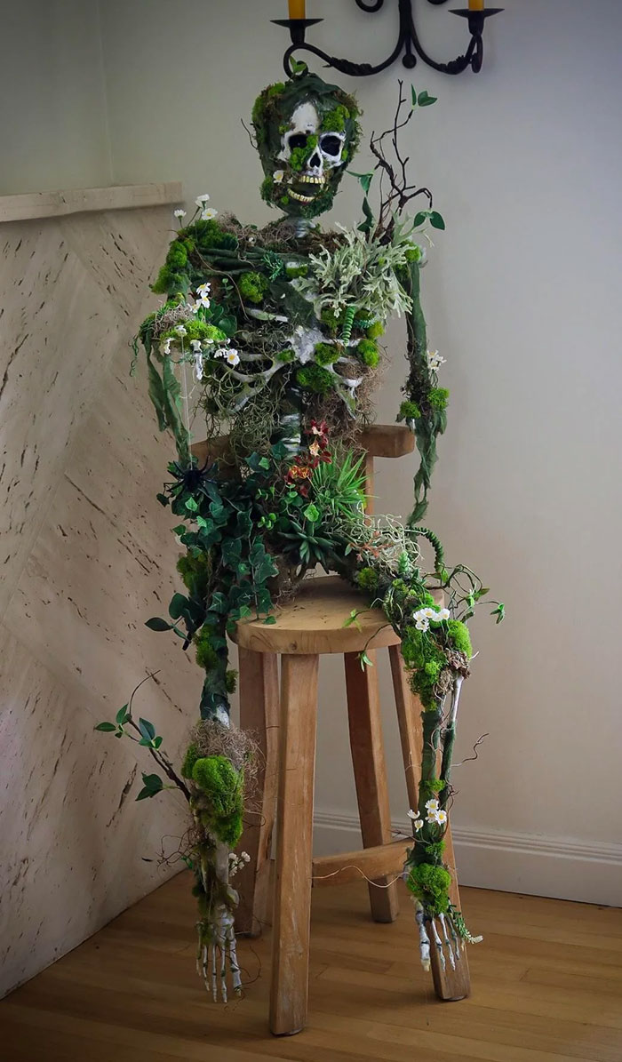 Creepy handmade craft resembling a skeleton adorned with moss and leaves, sitting on a wooden stool in a dim room.