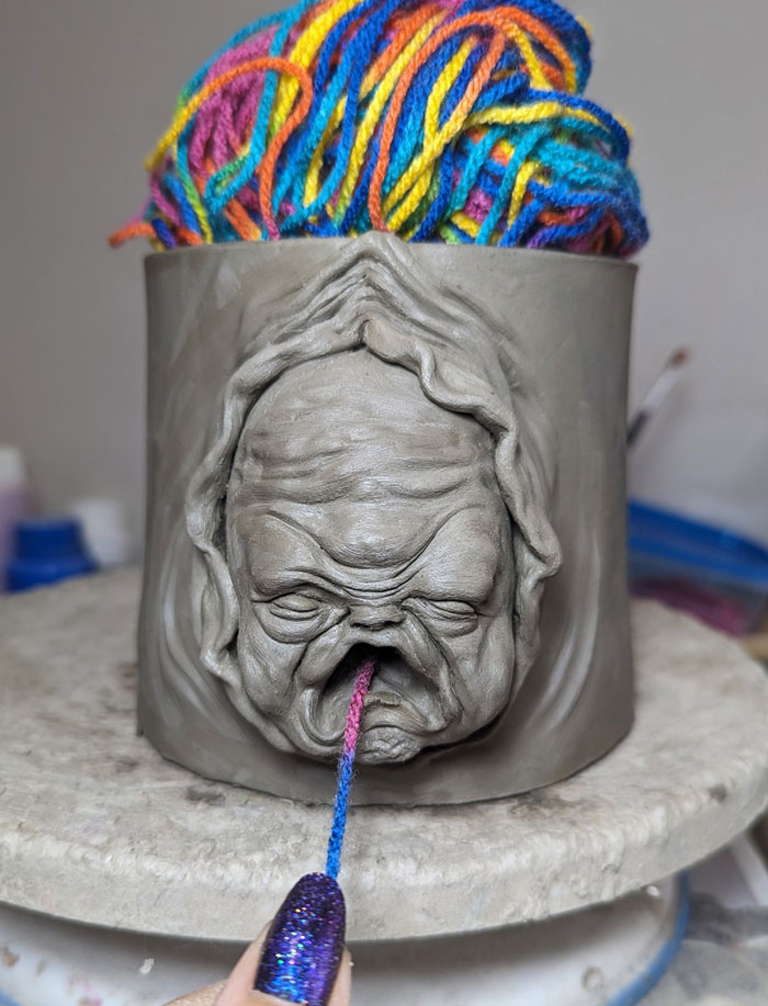 A creepy handmade clay face on a yarn holder, with colorful yarn emerging from its mouth.