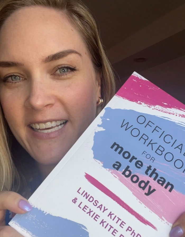 A woman holding a self-help workbook, related to body positivity, smiling at the camera. A woman holding a self-help workbook, related to body positivity, smiling at the camera.